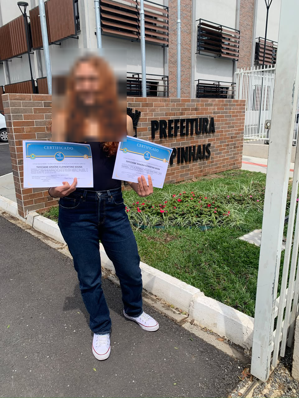 Thyciane segurando certificados em frente à Prefeitura de Pinhais, com o rosto pixelado